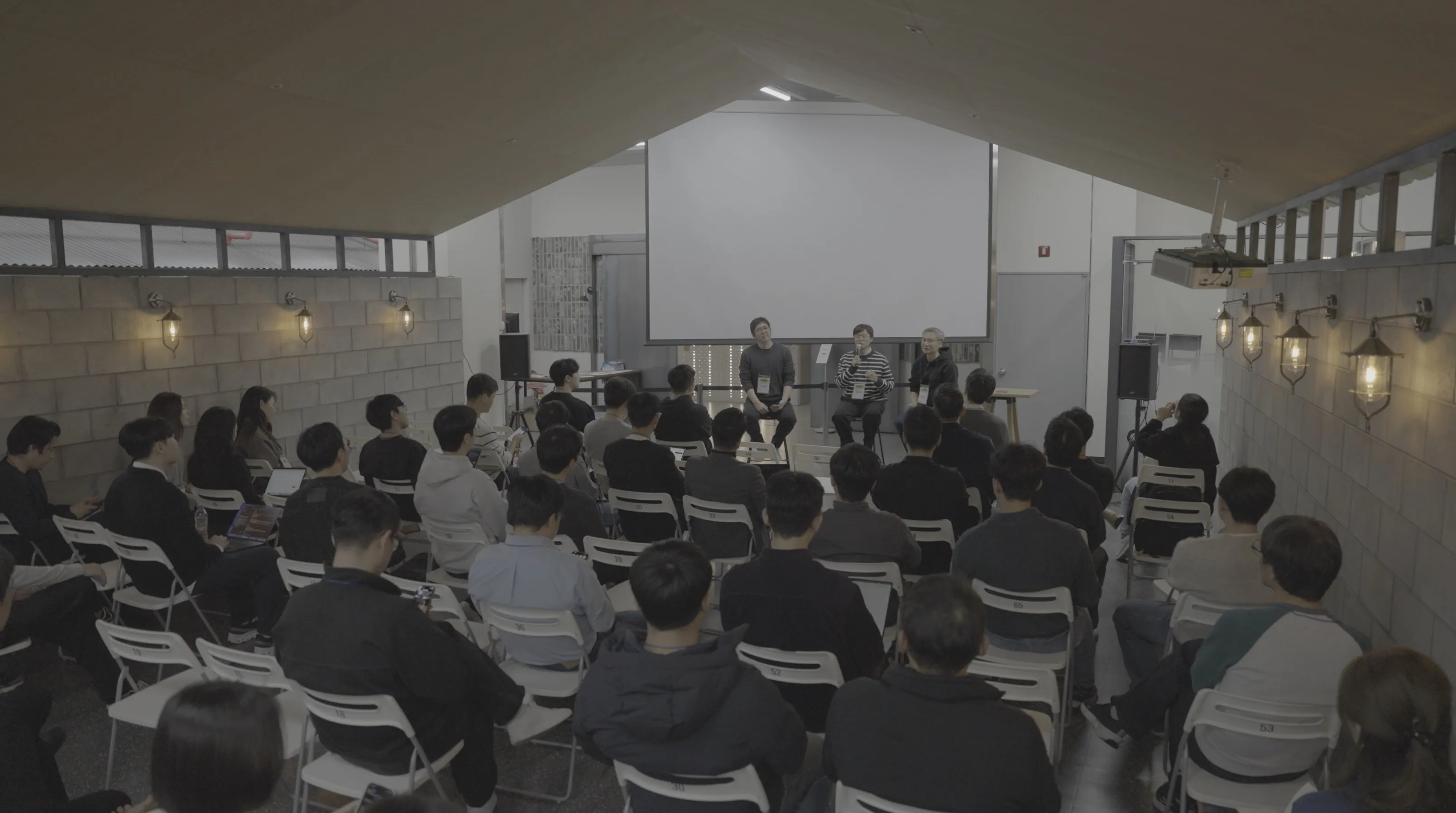 어두운 공간에서 청중 약 40명이 의자에 앉아 앞쪽의 패널 3인을 바라보고 있는 도망자 연합 패널 토의 전경. 스크린과 조명이 설치된 무대 앞